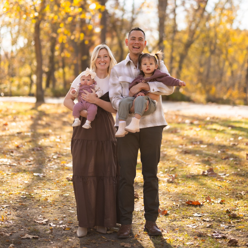 Family of four smiling outdoors: two parents holding young children, autumn foliage in background.