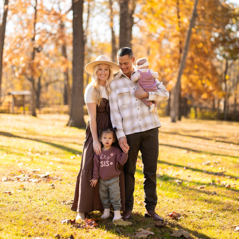 Family of four smiling outdoors in autumn: parents, toddler, baby.