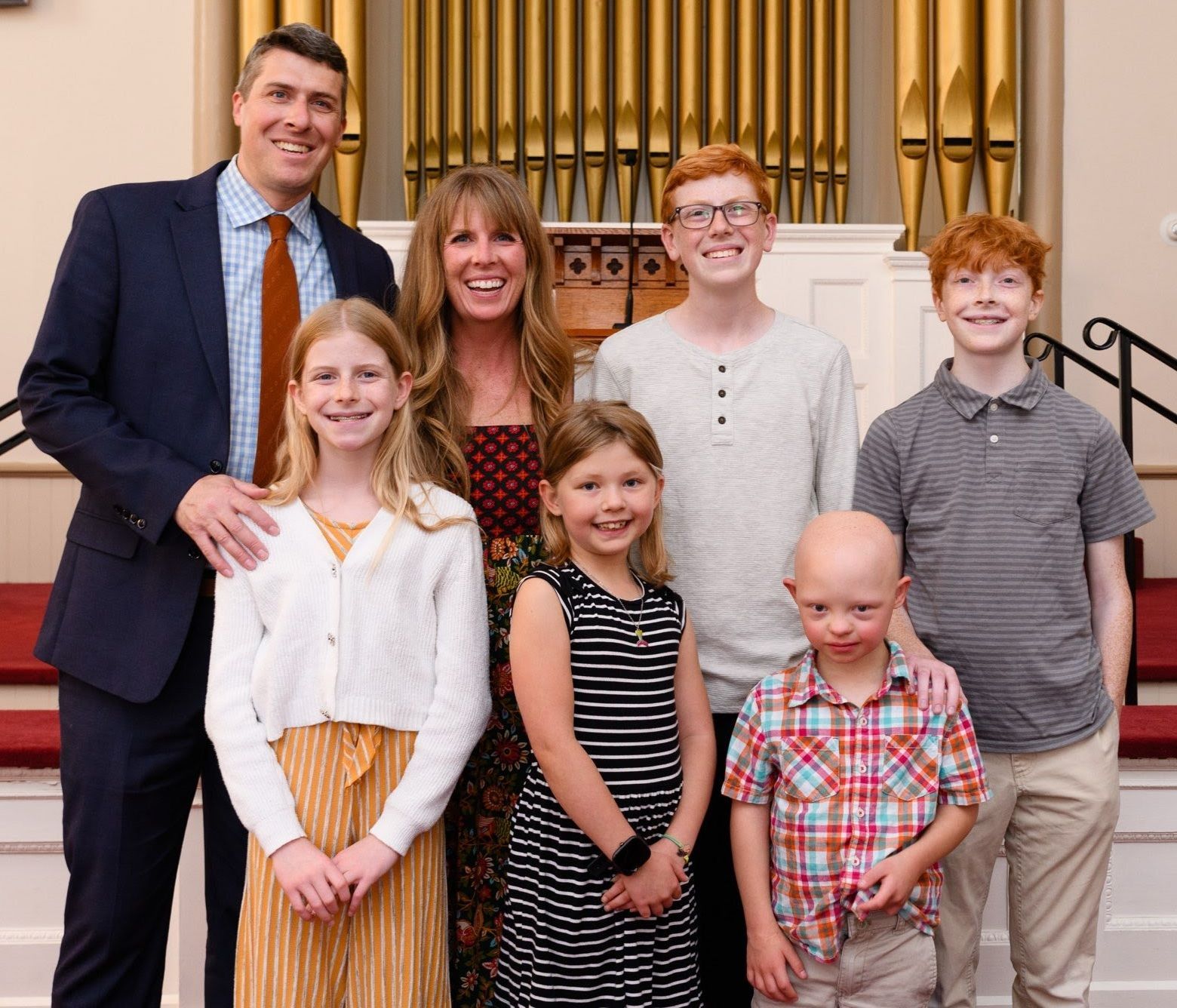 Family of seven poses in front of an organ. Smiling. Man in suit, woman in floral dress, children.