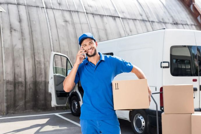Repartidor con uniforme azul que lleva una caja junto a una furgoneta blanca, hablando por teléfono.