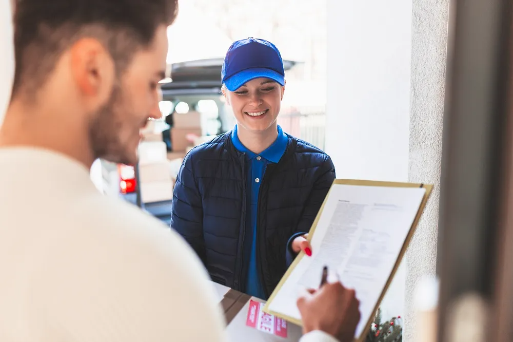 Una persona sonriente con gorra azul entrega un portapapeles en un mostrador de servicio.