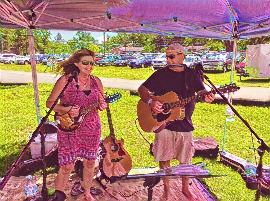 A man and a woman are playing guitars and singing into microphones under a tent.