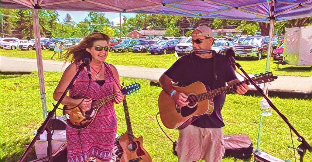 A man and a woman are playing guitars and singing into microphones under a tent.