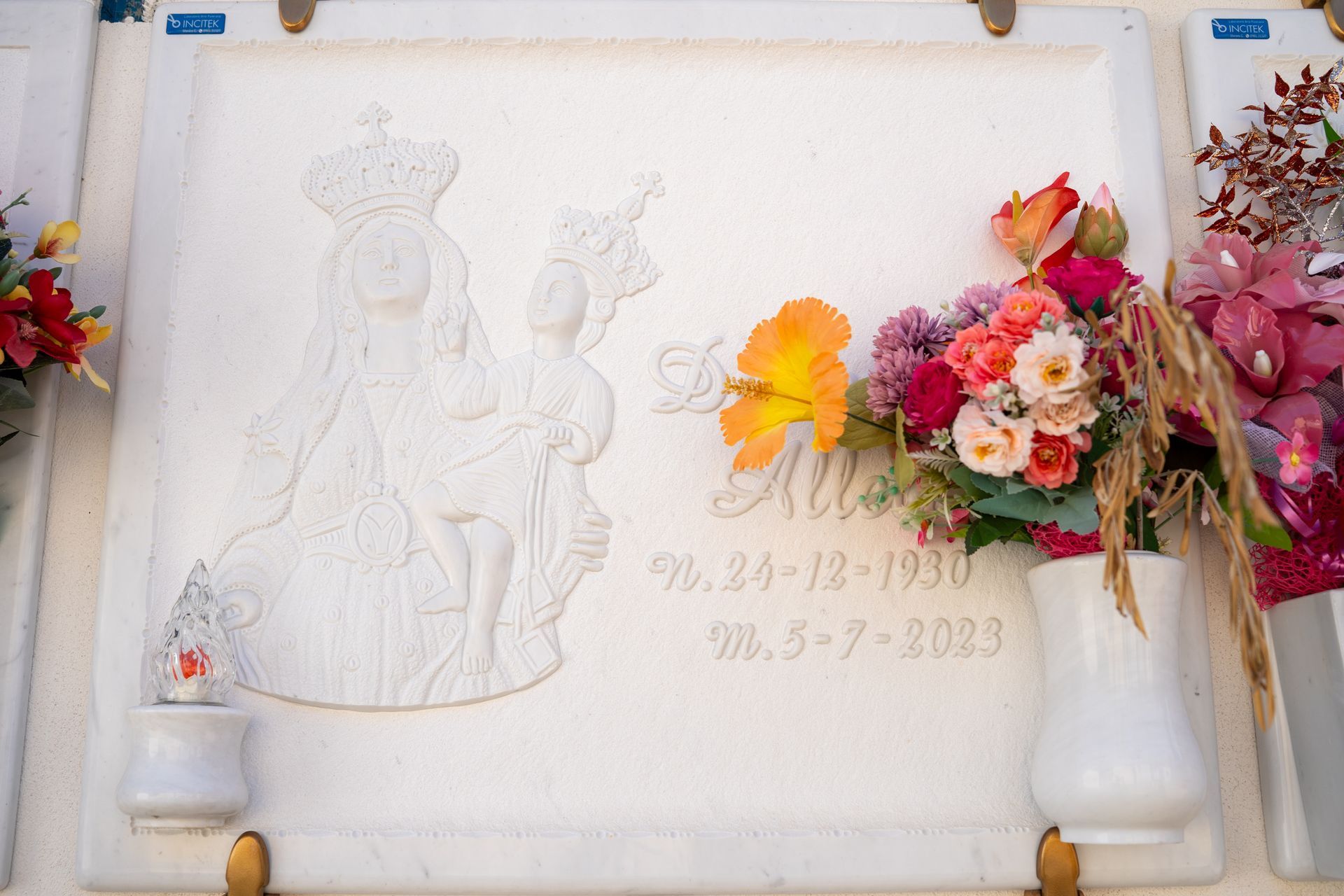 Lapide in marmo bianco con figura religiosa scolpita e fiori; date: dal 24-12-1930 al 20-7-2023.
