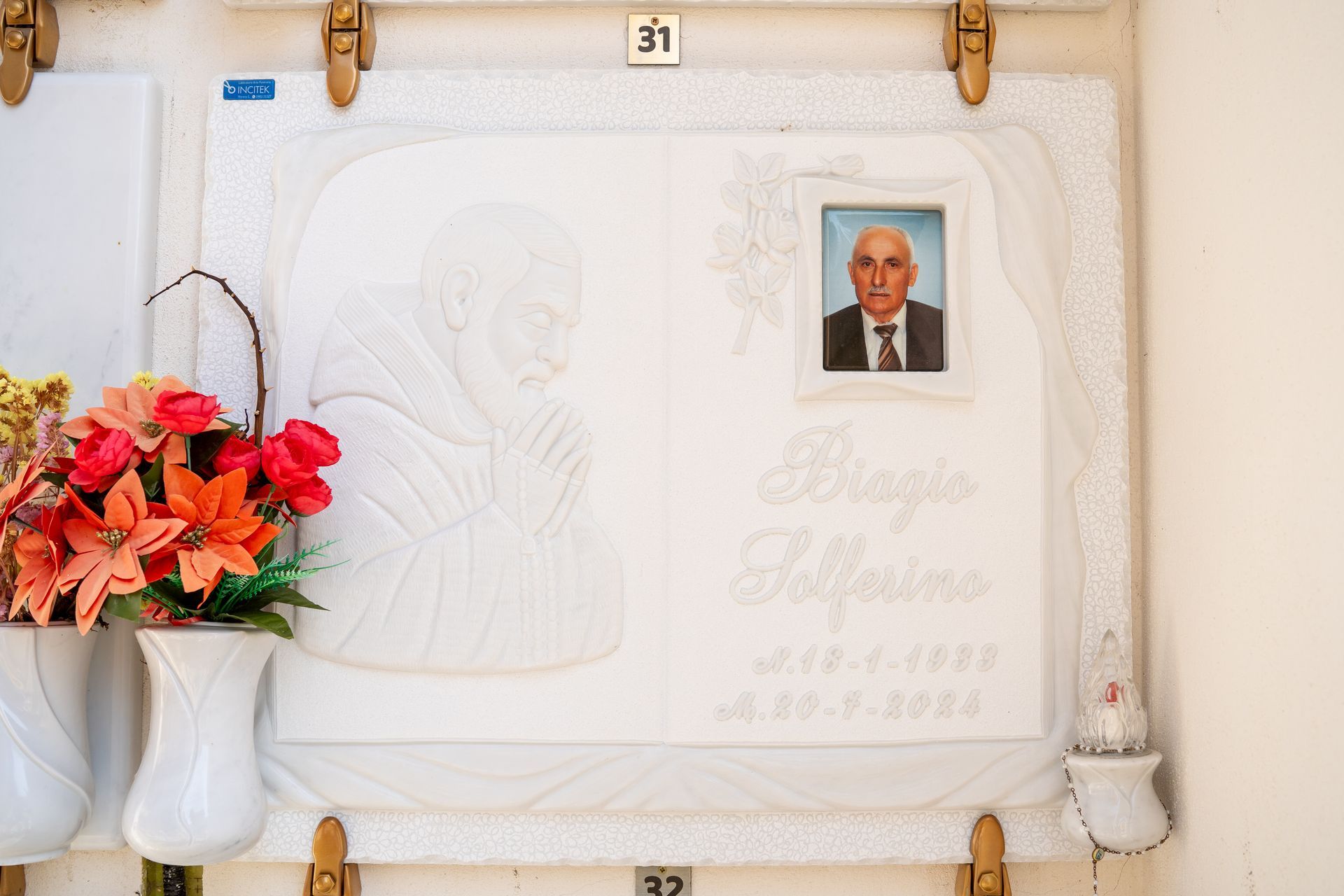 Lapide in marmo bianco con la foto di un uomo e l'immagine incisa di una figura religiosa, ornata di fiori.