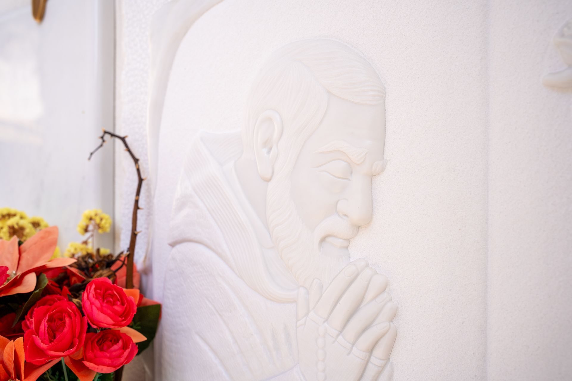 Rilievo in marmo bianco raffigurante un uomo con la barba e le mani giunte in preghiera, con fiori in primo piano.