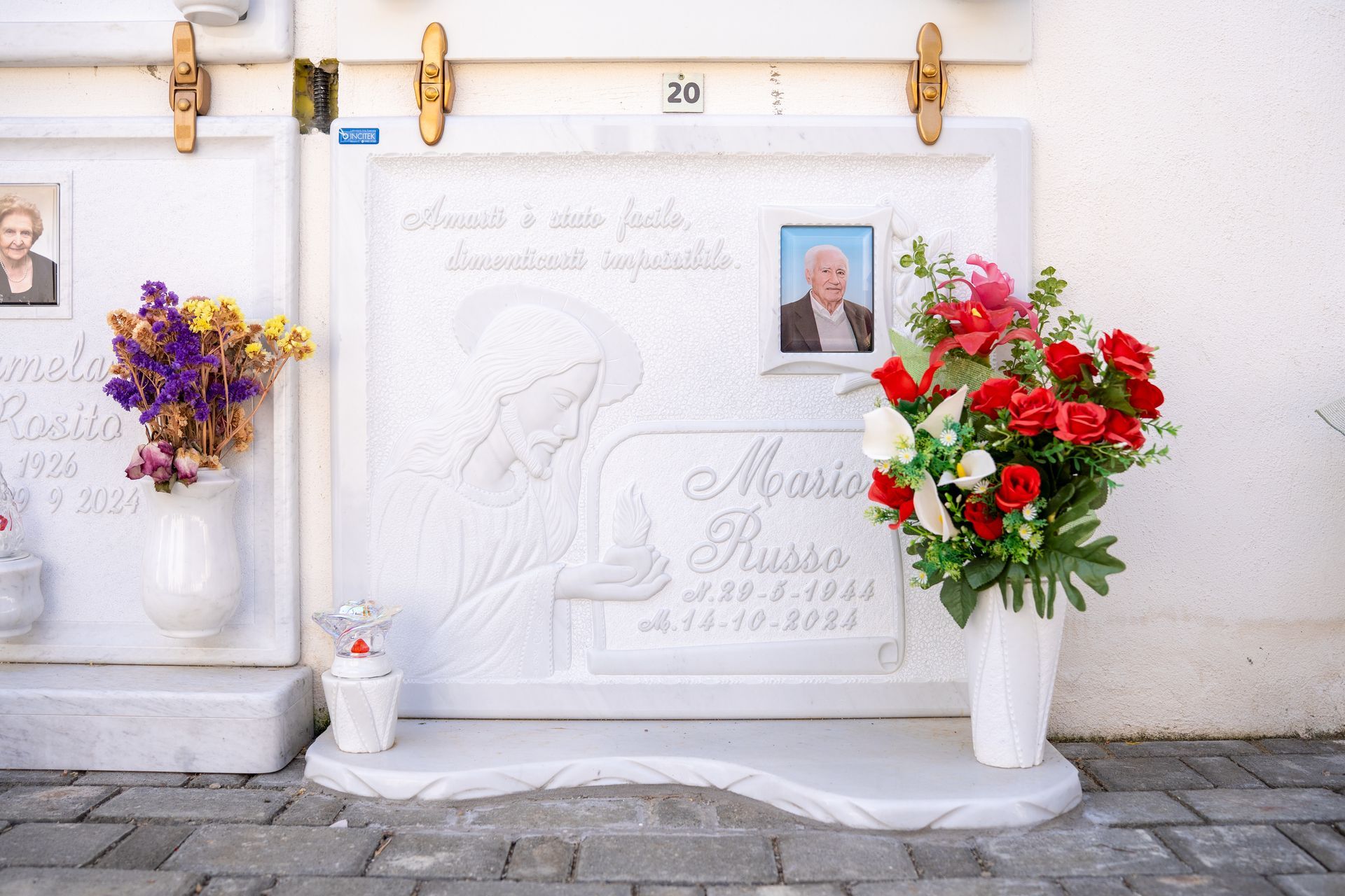 Lapide con composizioni floreali e un ritratto. Immagine scolpita di una persona in lutto. Fiori rossi e bianchi e un'altra con fiori viola.