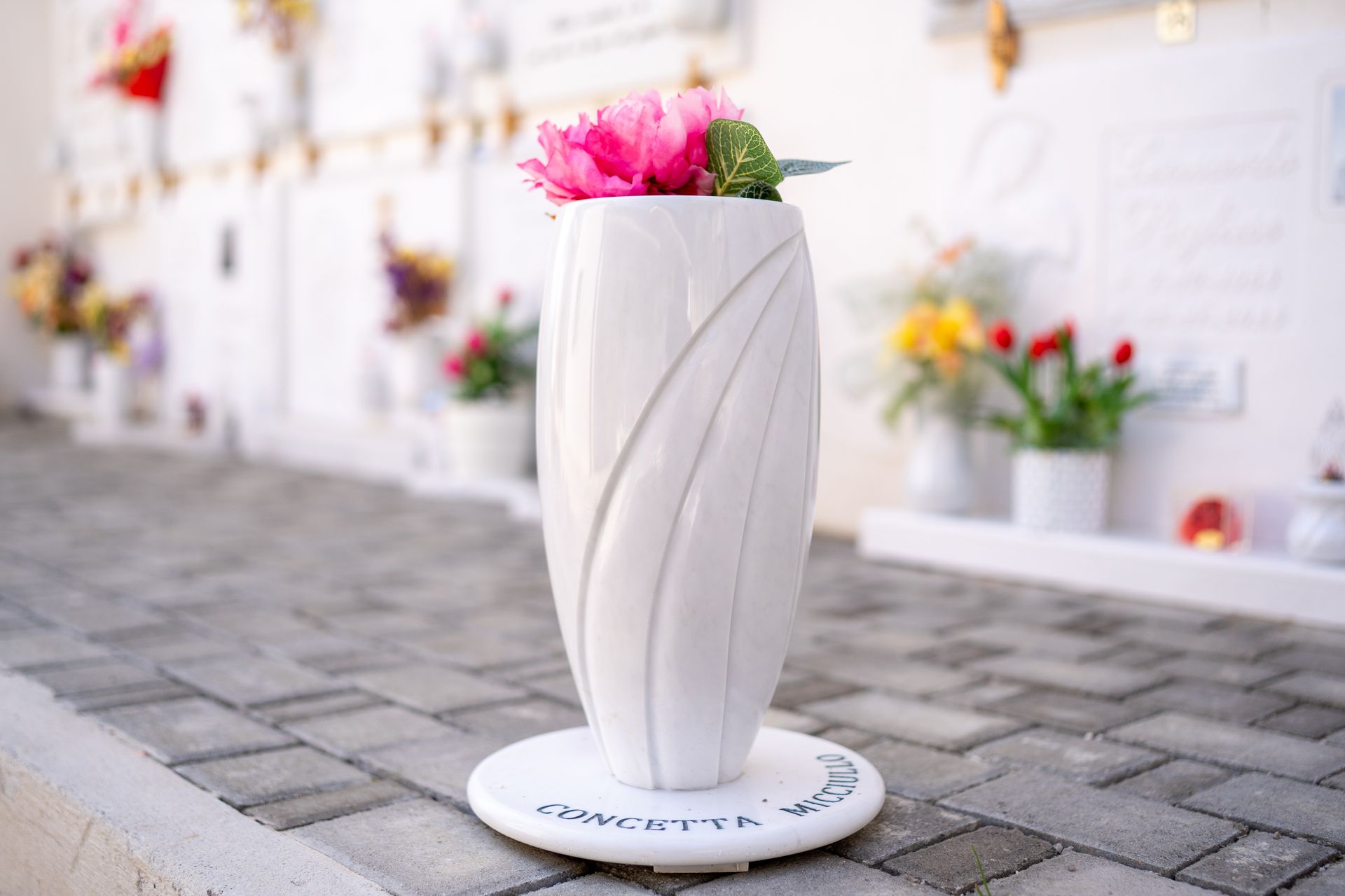 Vaso bianco con fiori rosa su una tomba, con una base circolare bianca su un pavimento in mattoni grigi, altre tombe sullo sfondo.