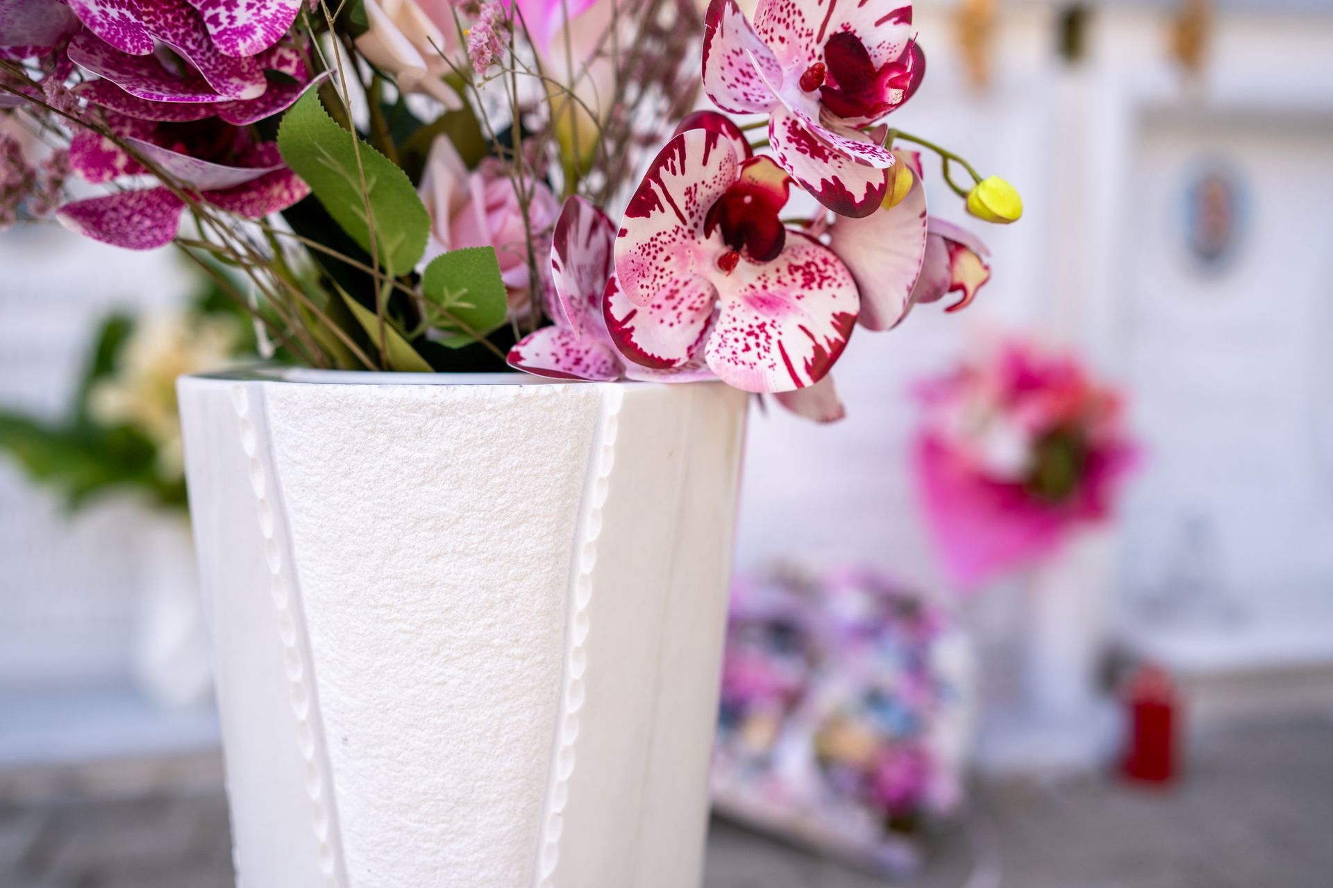 Primo piano di un vaso bianco geometrico pieno di orchidee rosa e viola, sullo sfondo un cimitero.
