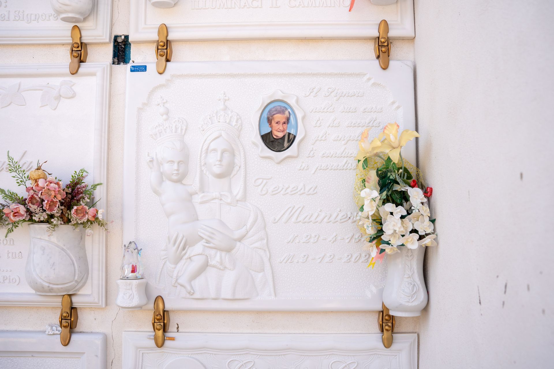 Tomba in marmo bianco con ritratto e scultura in rilievo di Maria che tiene in braccio Gesù, fiori e decorazioni dorate.
