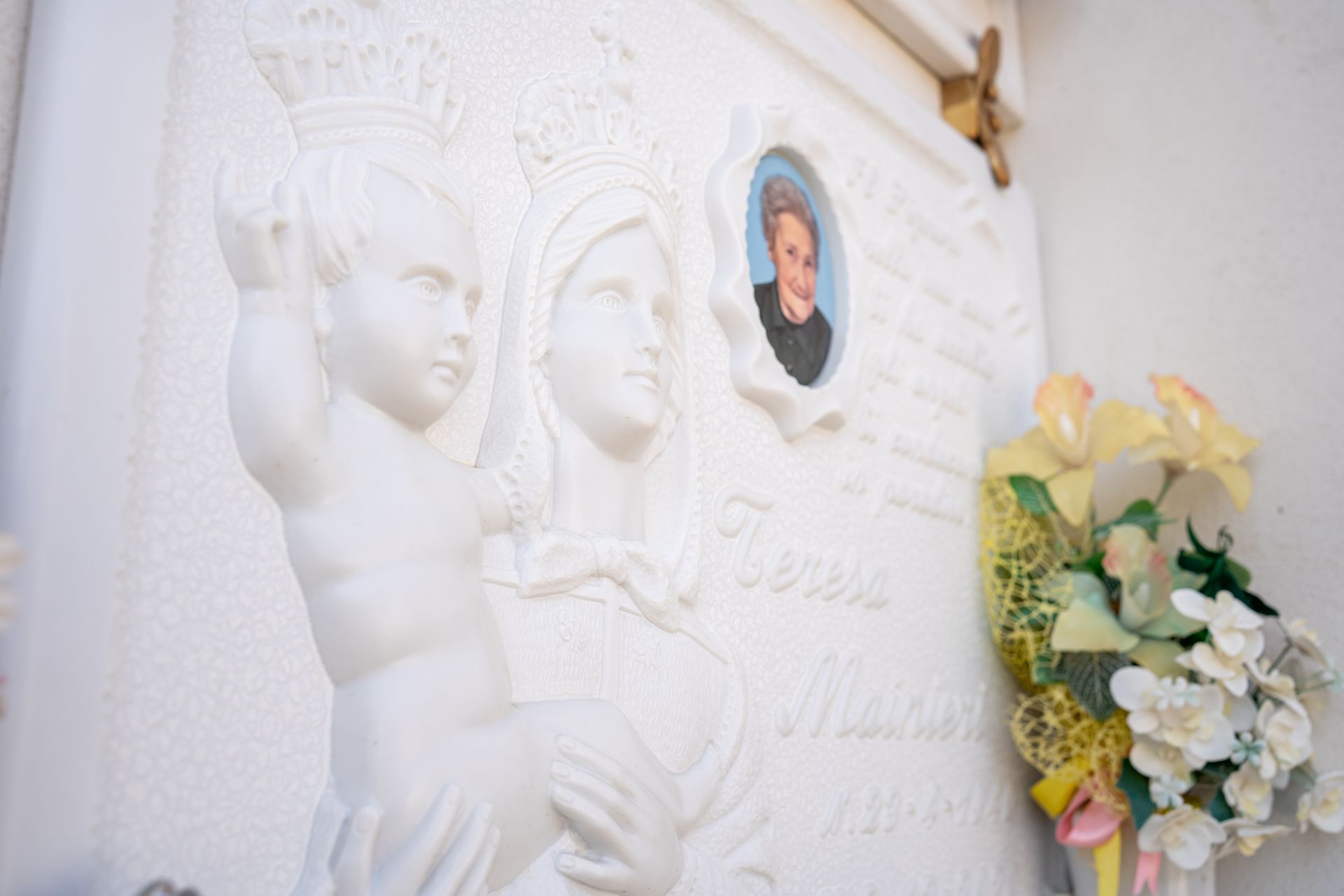 Tomba bianca decorata con una statua della Vergine Maria con il bambino, una foto e dei fiori.