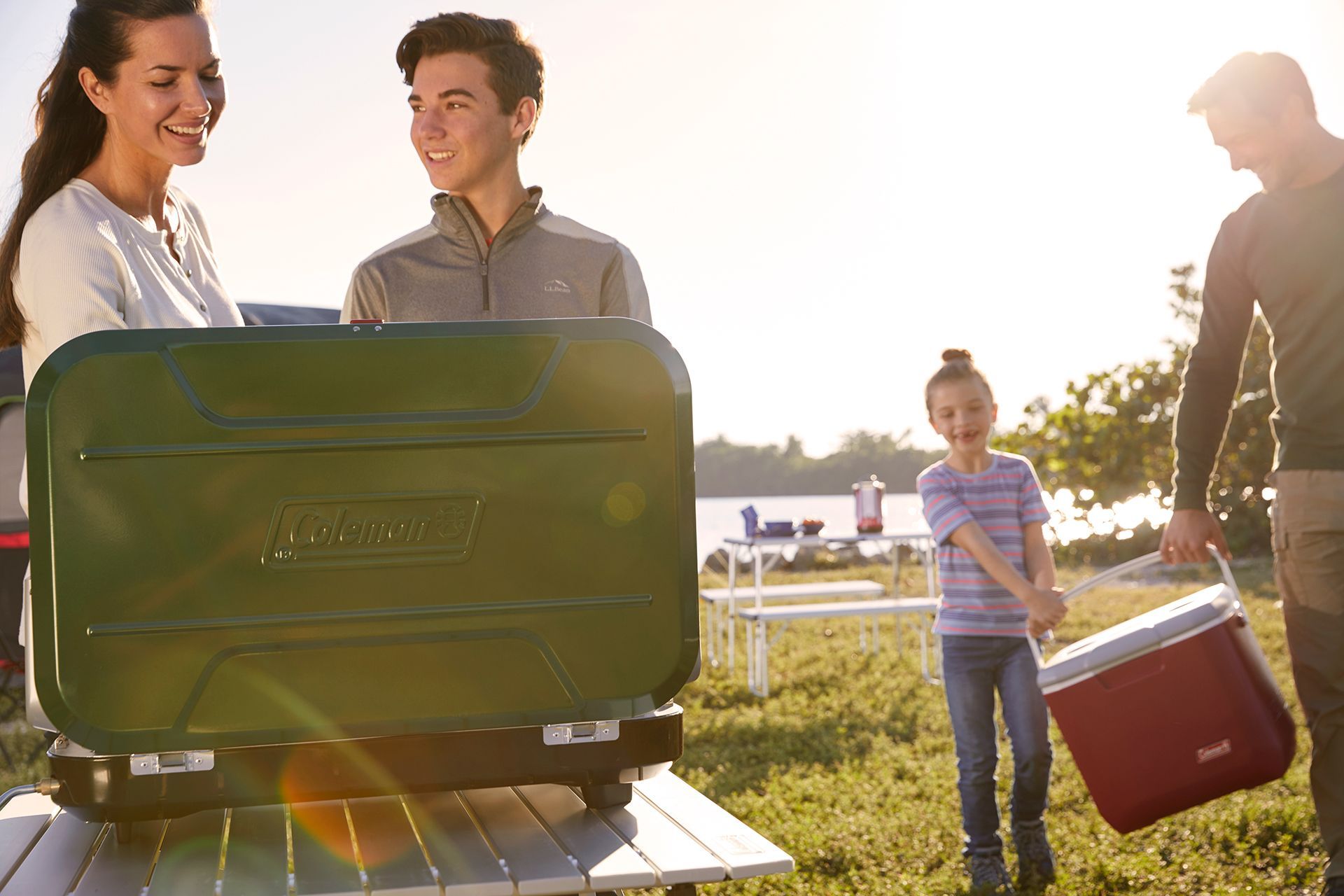 A couple is standing next to each other cooking on a Coleman grill and a father and daughter are carrying a Coleman cooler.