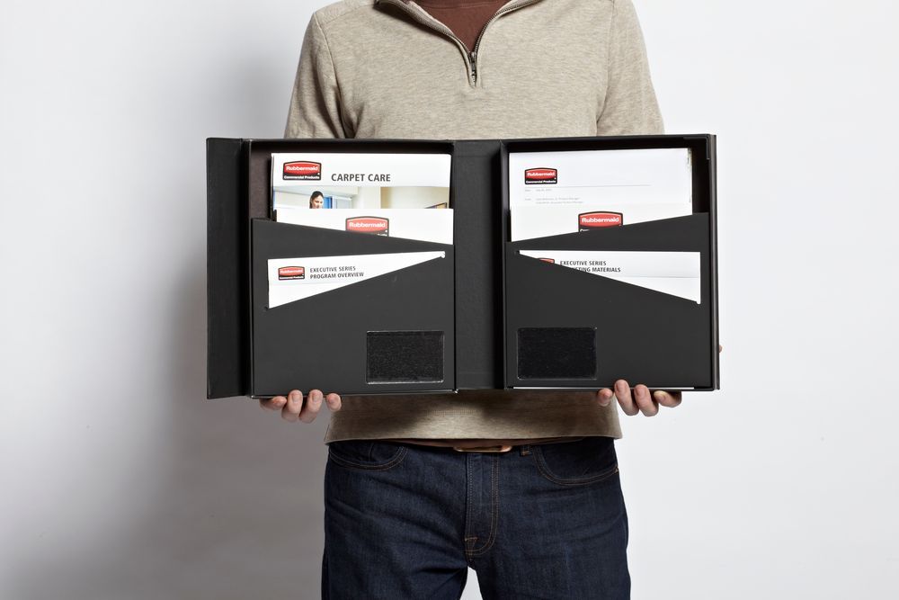 A man is holding a black folder with with all the launch material for a Rubbermaid commercial products launch