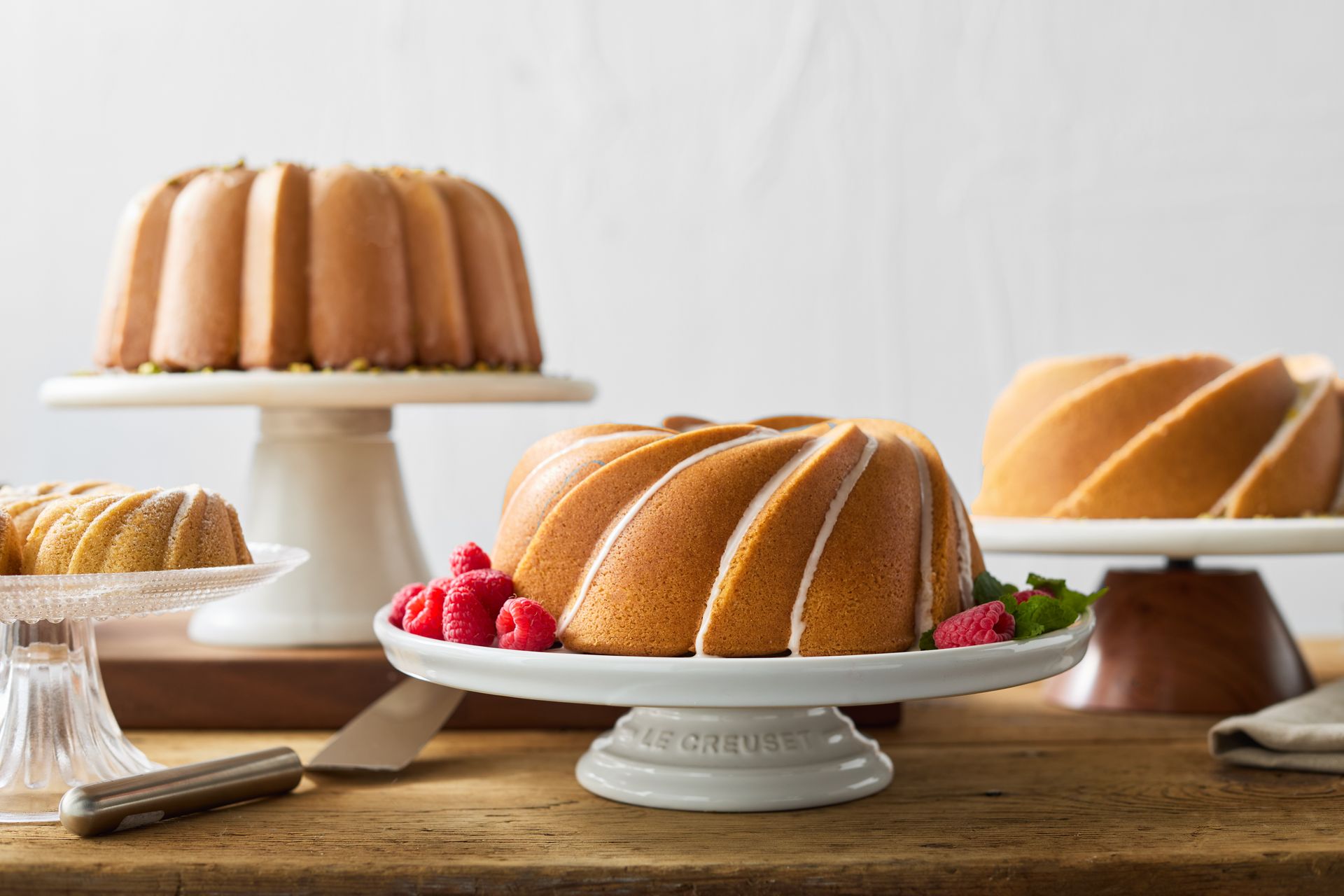 Three bundt cakes are sitting on cake stands on top of a wooden table.