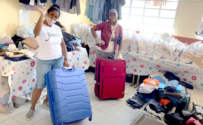 Two women are standing next to suitcases in a room filled with clothes received from JR