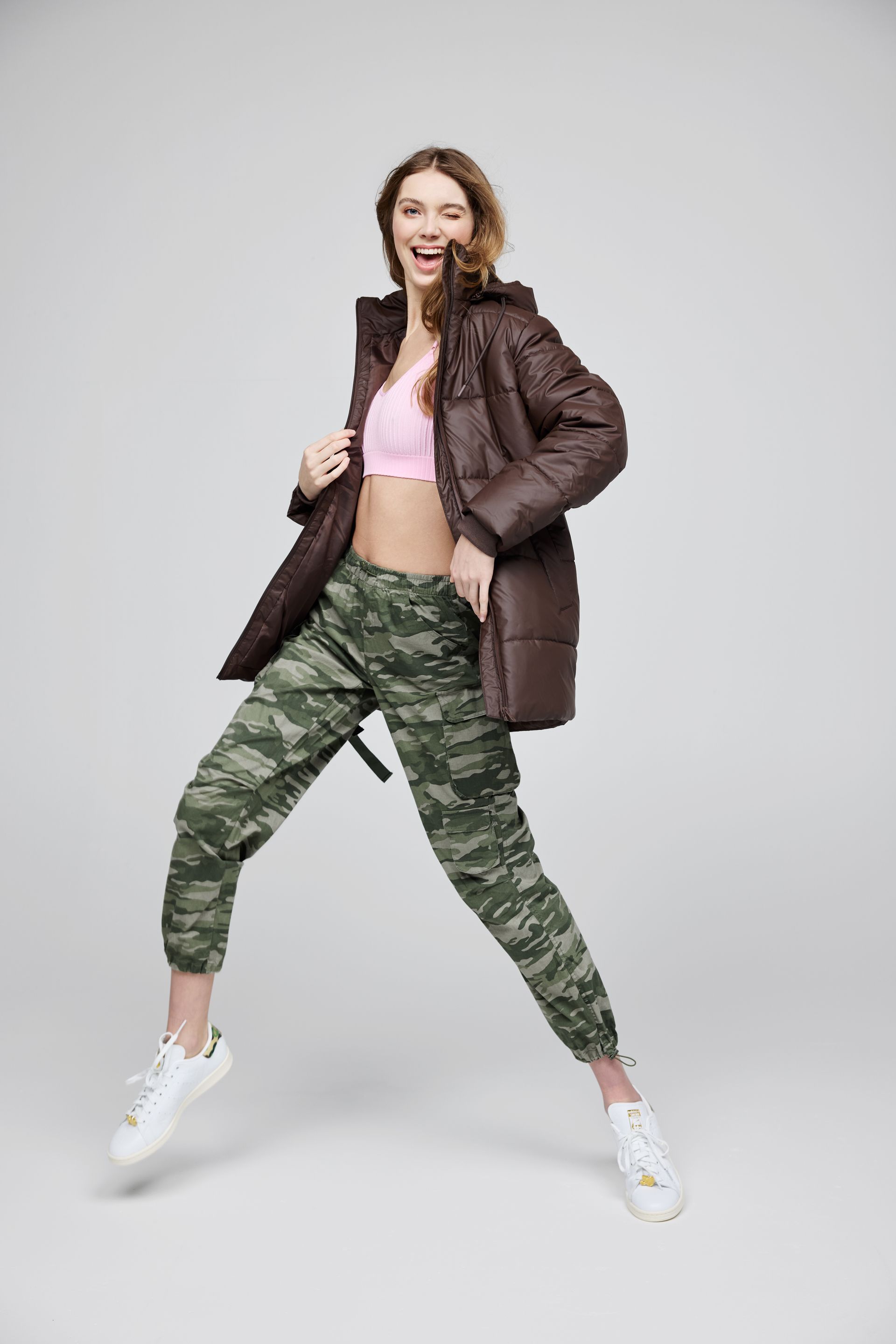 A woman in a brown jacket and camo pants is jumping in the air.