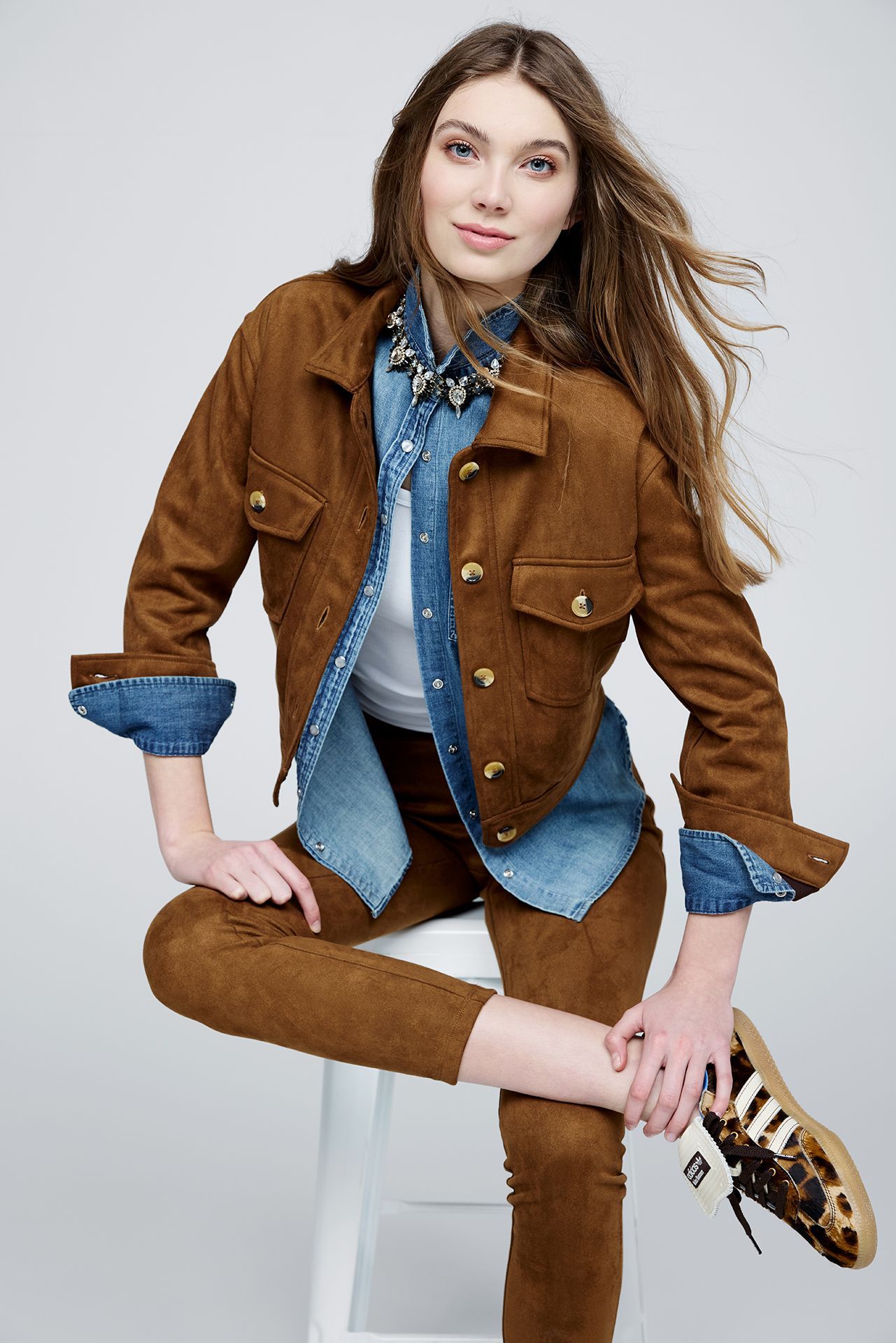 A woman is sitting on a stool wearing a brown jacket and denim shirt.