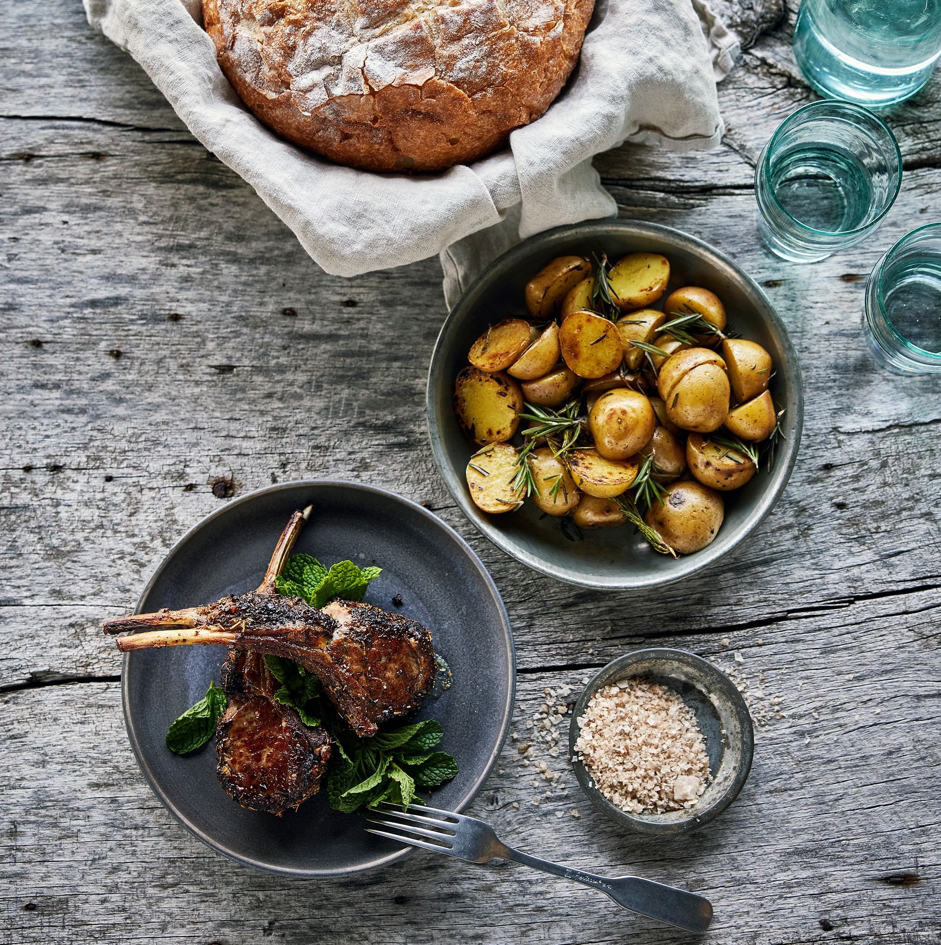 A wooden table topped with bowls of food including lamb chops and rosemary potatoes.