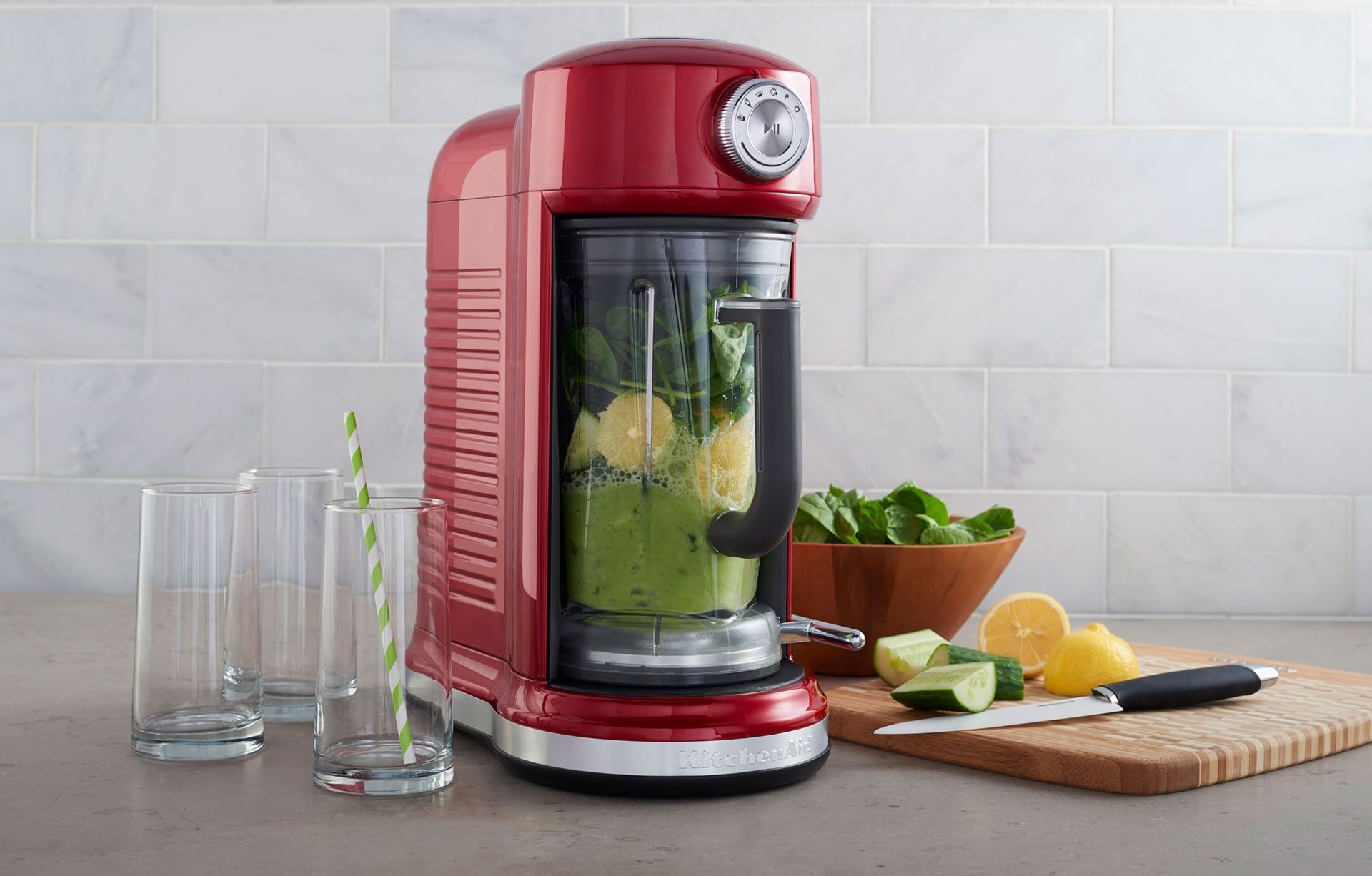 A red KitchenAid blender is sitting on a counter next to a cutting board and glasses.