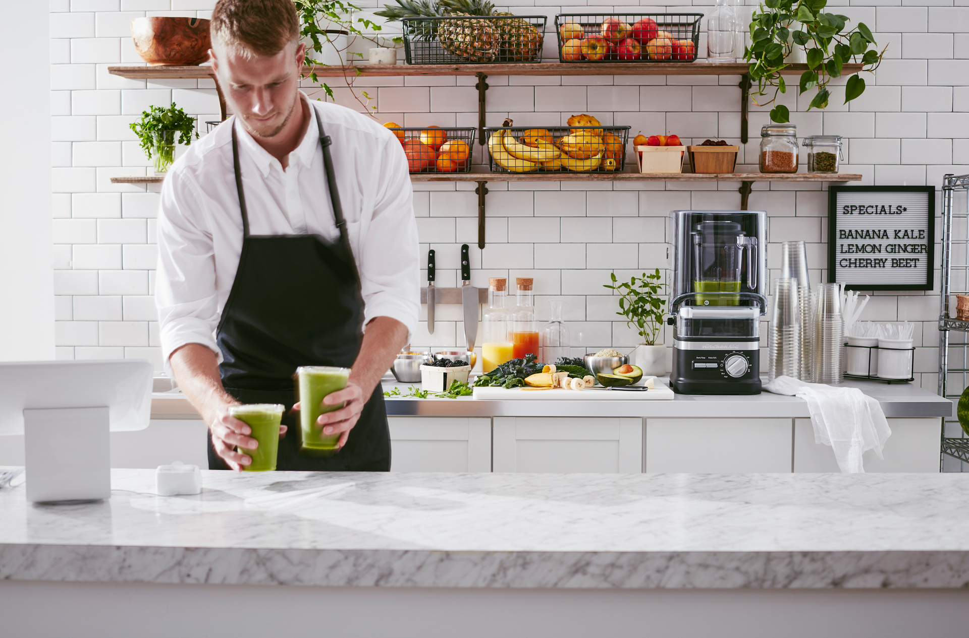 A man in an apron is making a green smoothie in a kitchen with a KitchenAid commercial blender in the background