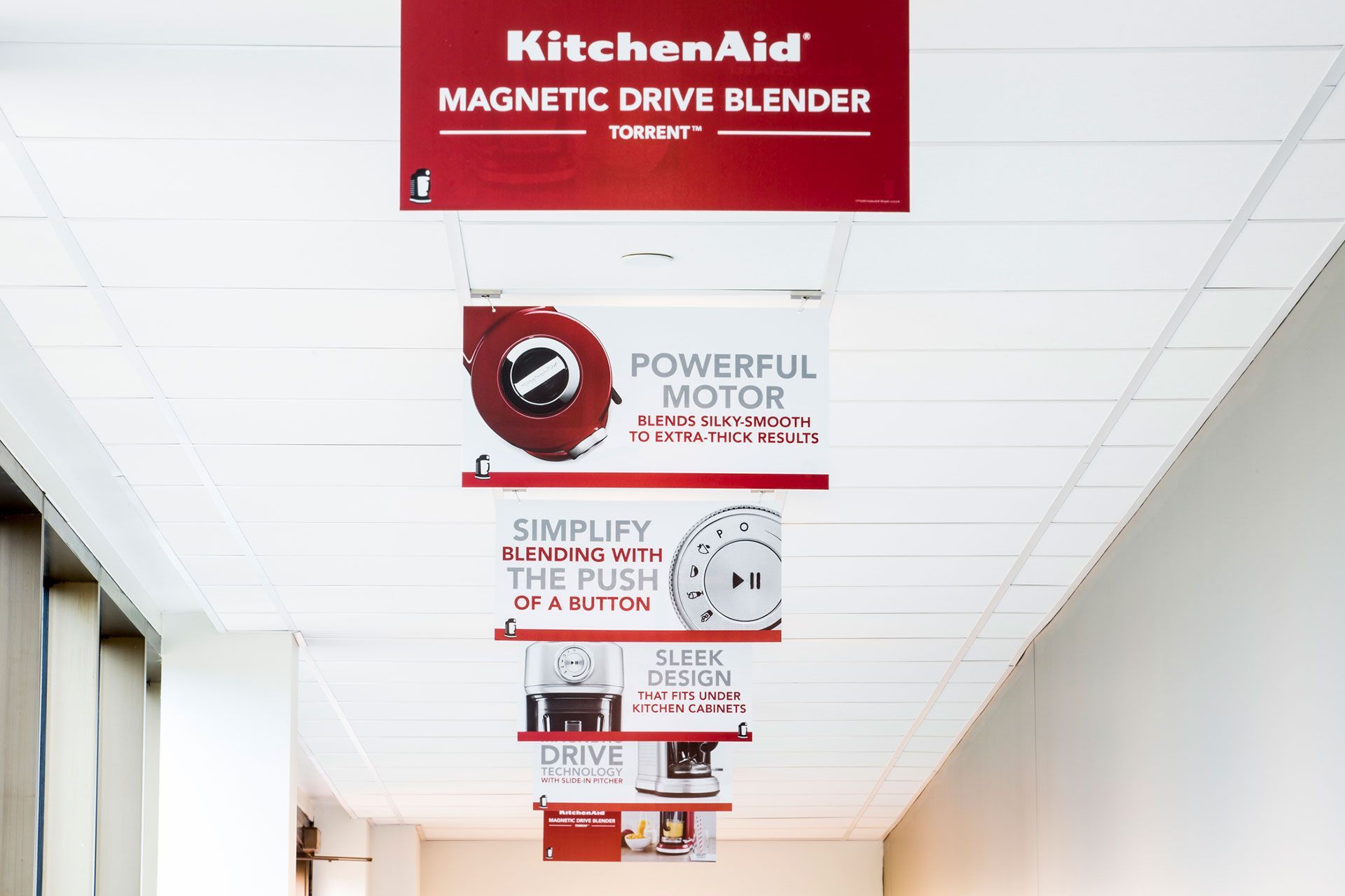 A launch sign showing a KitchenAid magnetic drive blender is hanging from the ceiling in a hallway of an office building