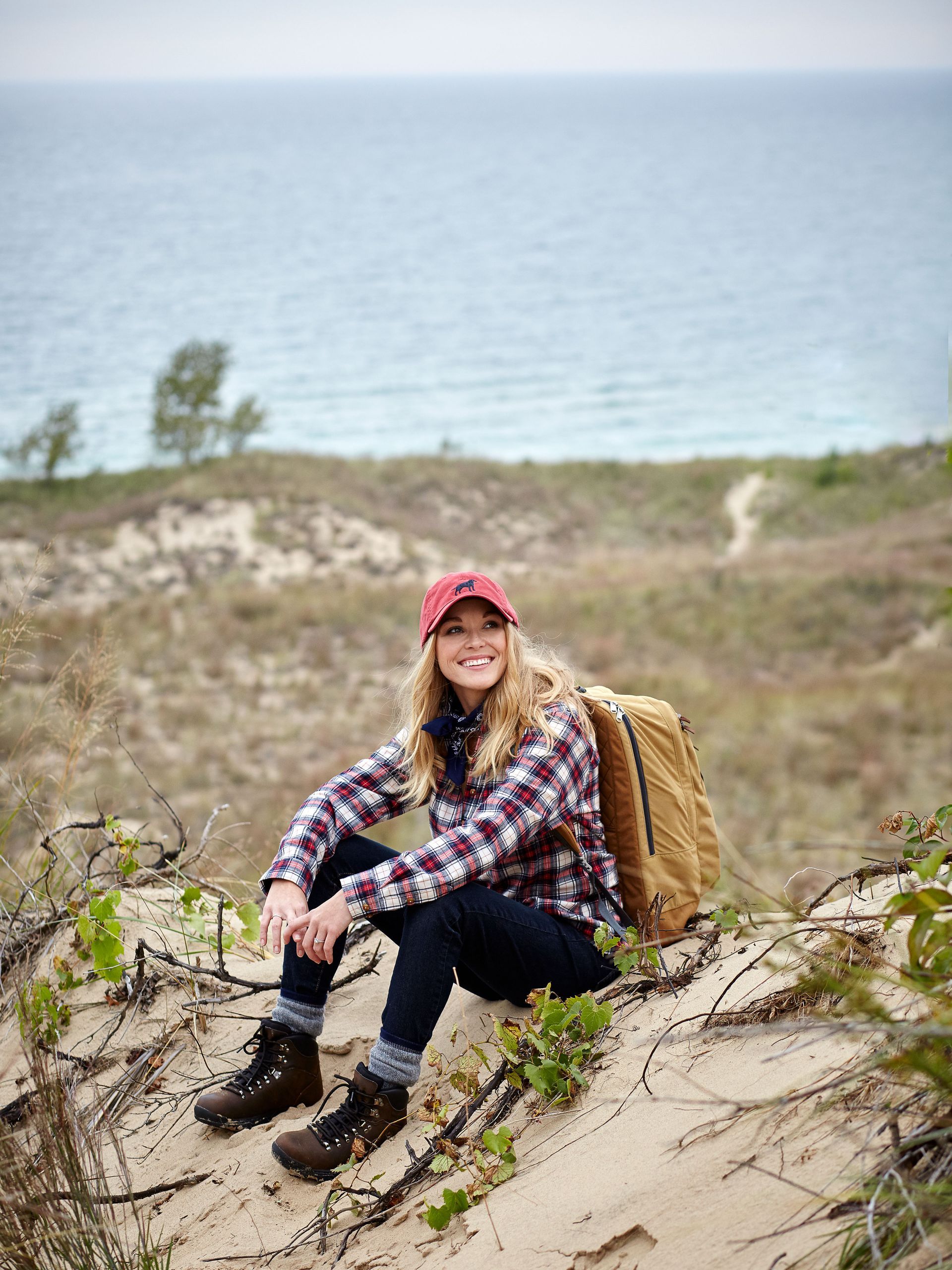A woman is sitting on top of a sand dune with a backpack.