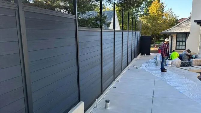 Backyard walkway beside a tall gray fence, with workers and supplies near a shed.
