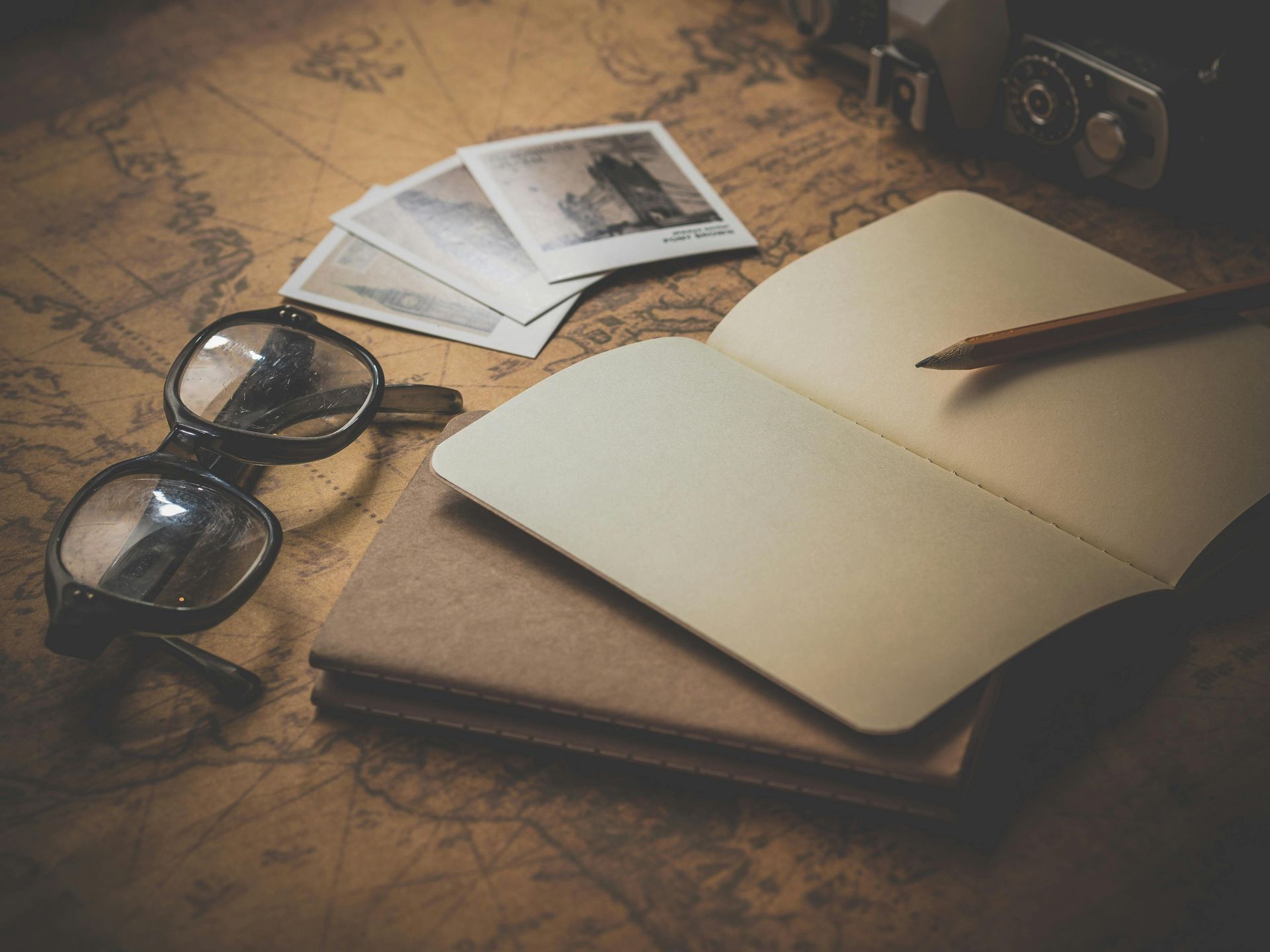 Open notebook, pencil, glasses, and photographs on a vintage map, suggesting travel and journaling.