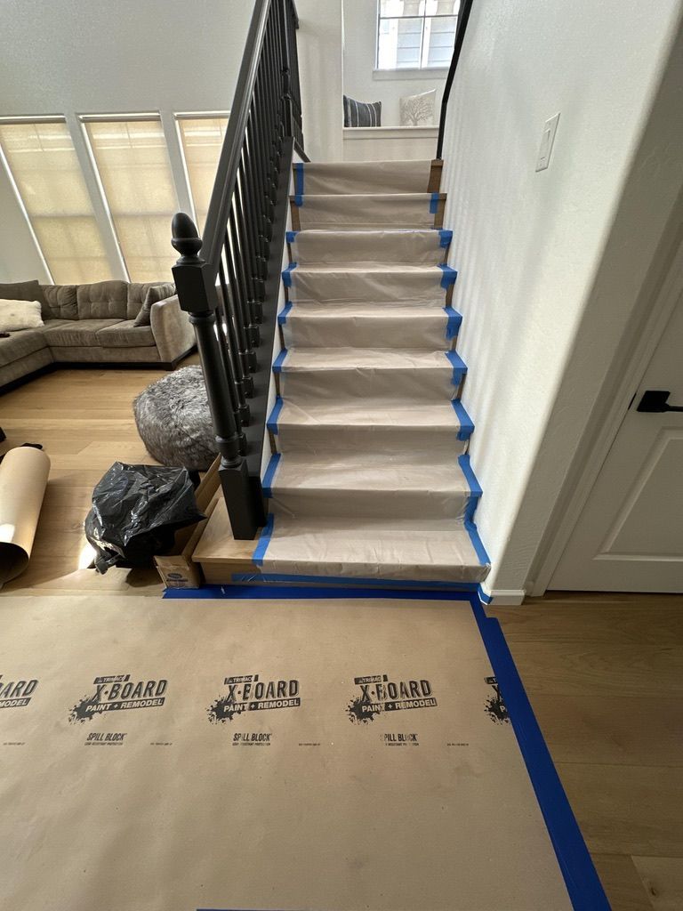 Staircase with protective covering; blue tape, black banister, and a living room in the background.