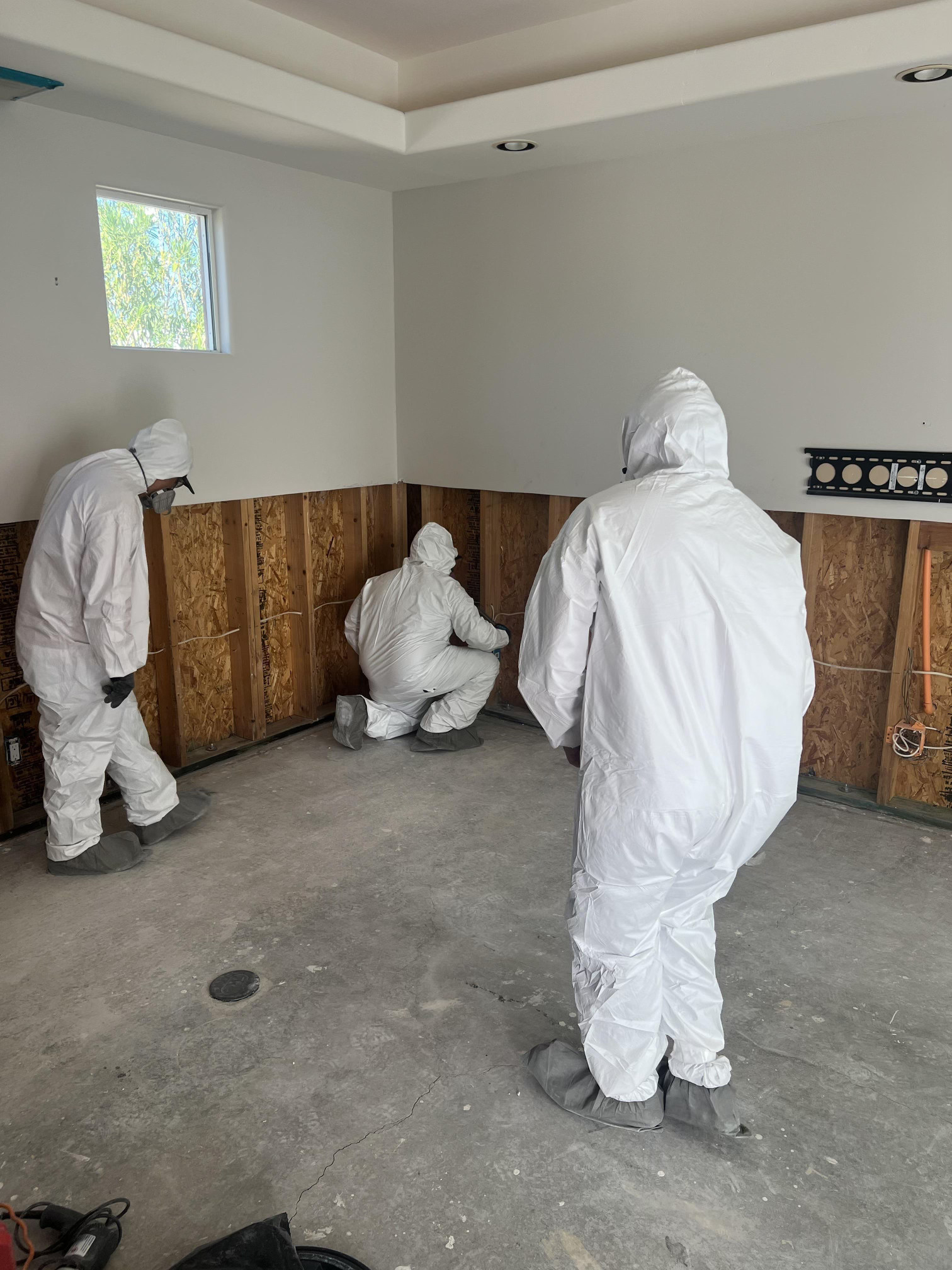 Three people in white hazmat suits are working in a room with exposed wall studs.