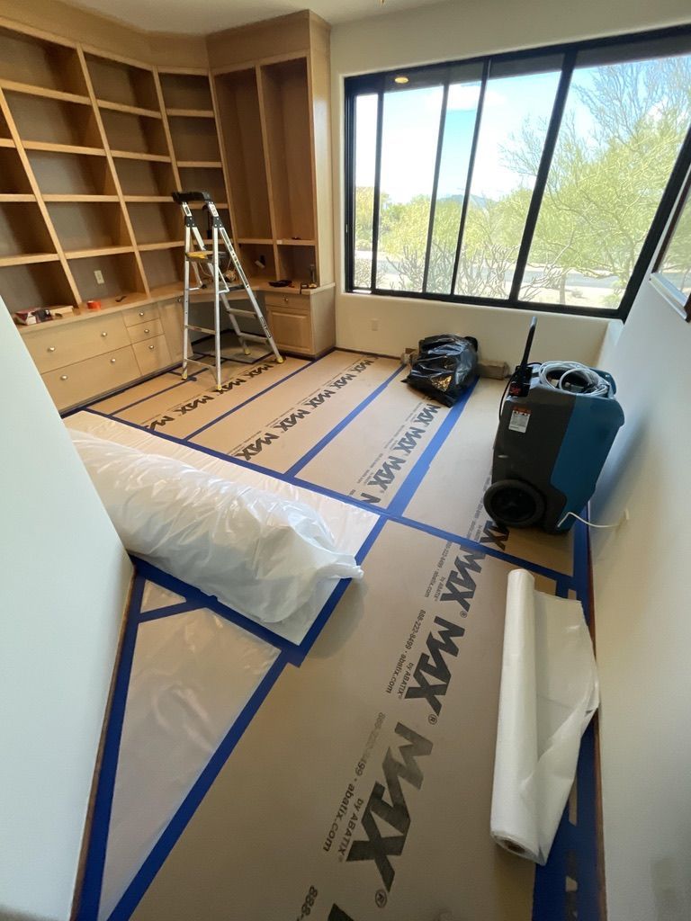 Room under construction with wooden shelving, blue tape, flooring protection, and equipment by a window.