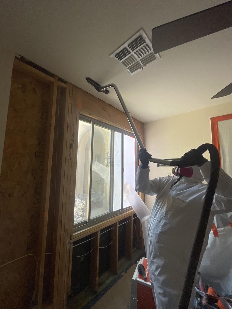 Person in hazmat suit vacuums near a window inside a room with exposed wall framing.