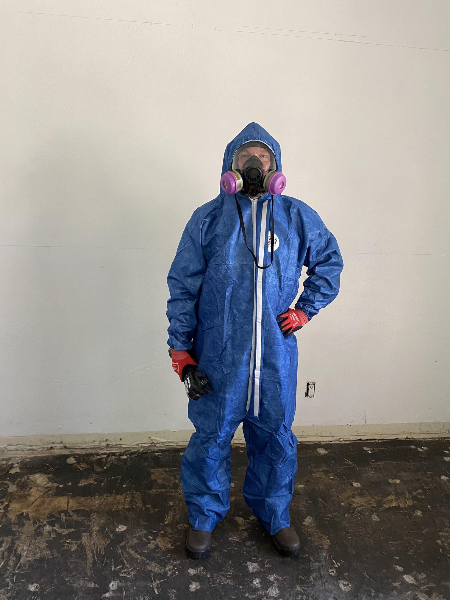 Person in blue hazmat suit, respirator, and gloves in a room, possibly cleaning or doing work.