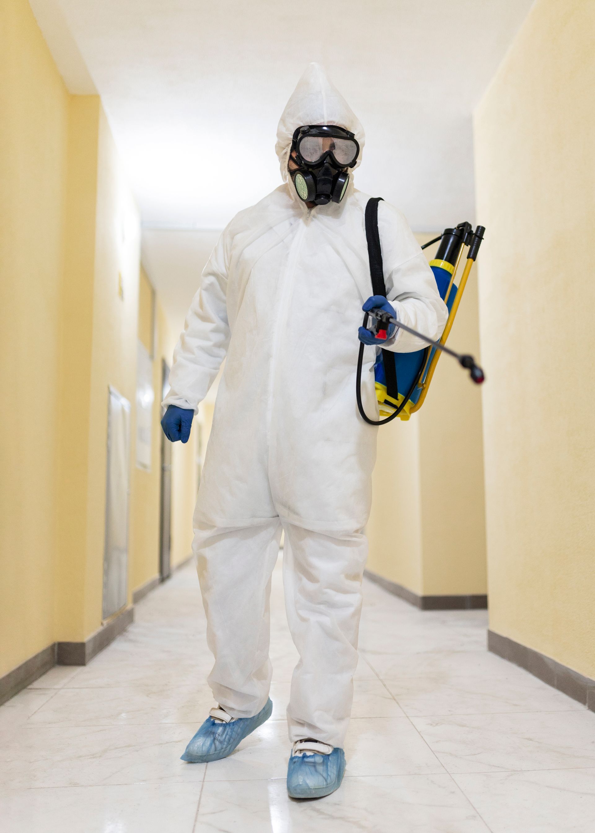 Person in white hazmat suit and respirator, holding a sprayer, in a hallway.
