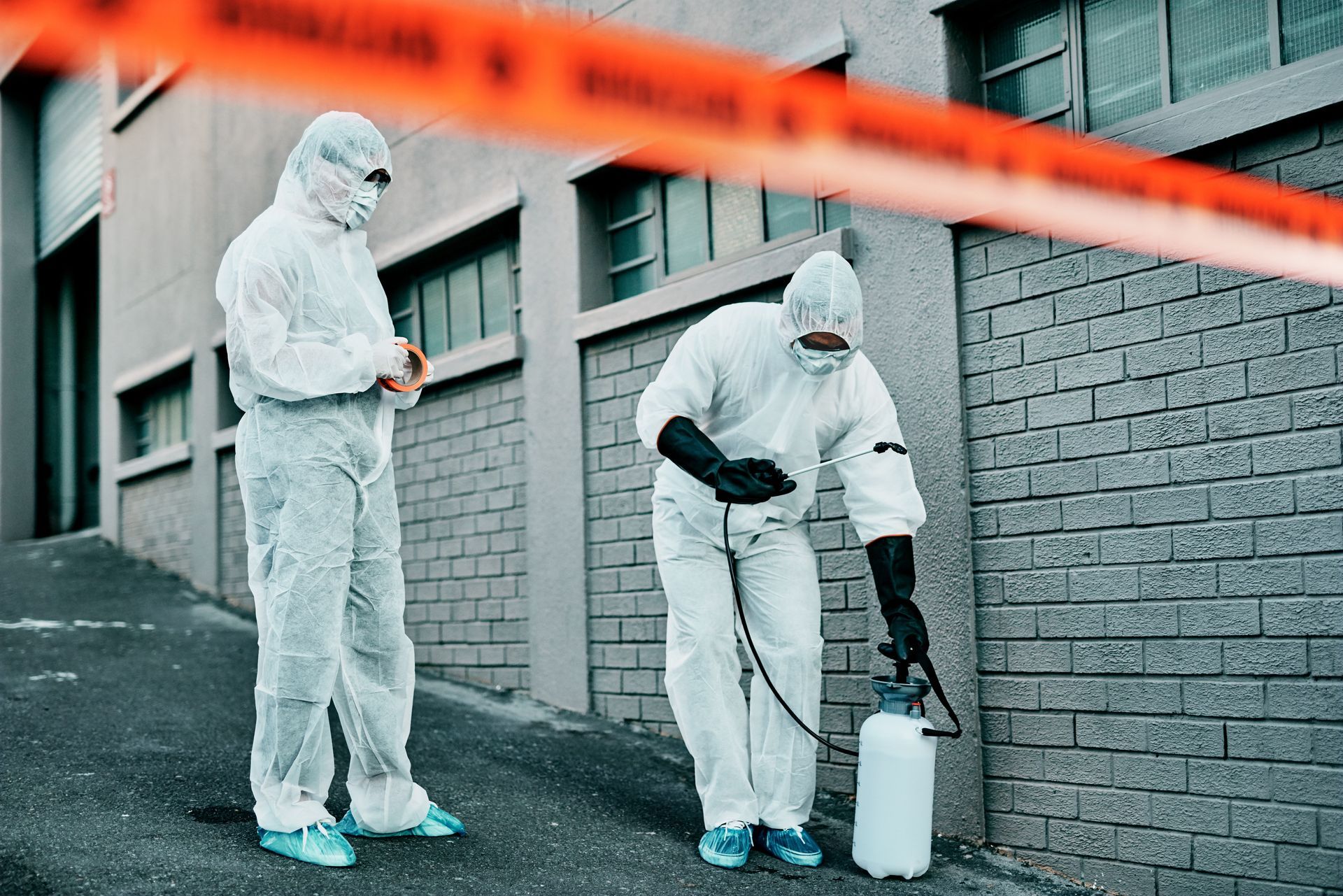 Two people in hazmat suits spraying a building exterior, blocked off by caution tape.