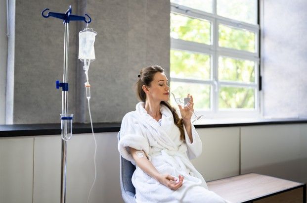 Woman in robe receiving IV drip, drinking water near a window.