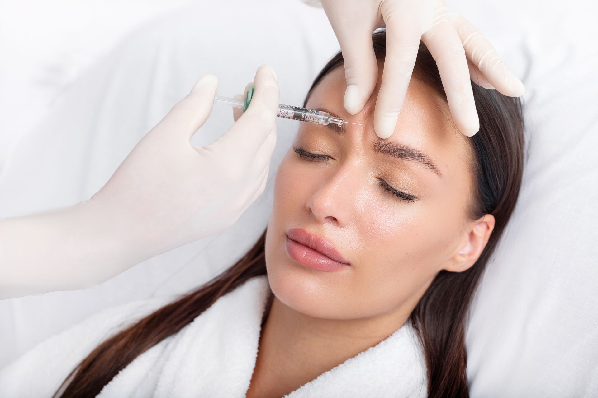 Woman receiving an injection between eyebrows with closed eyes, in a medical setting.