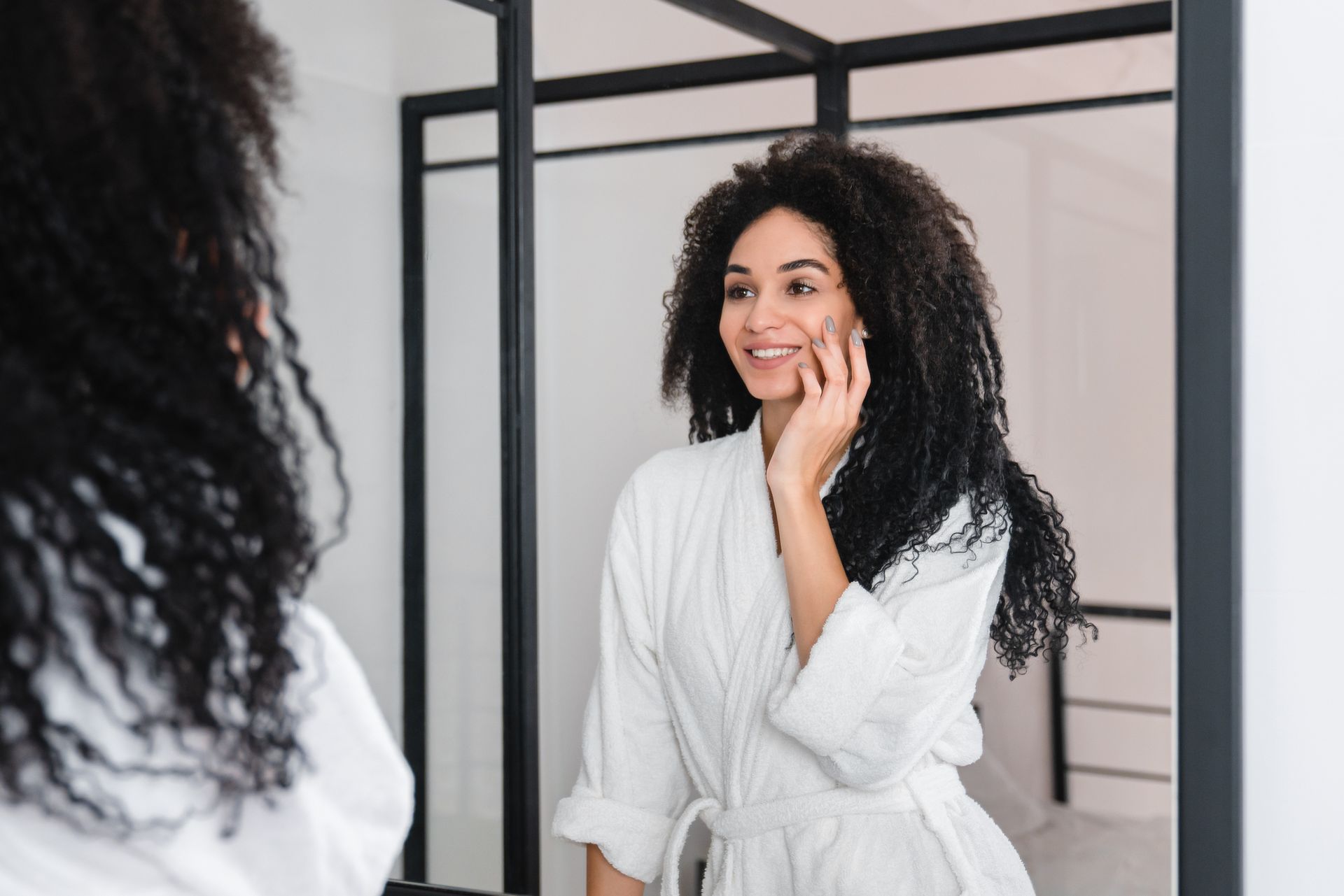 Woman in a white robe smiles while looking at herself in a mirror, touching her face.