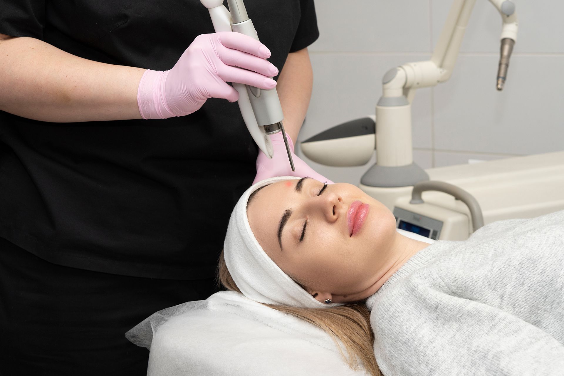 Woman receiving laser facial treatment in a clinic; a medical professional holds the device.