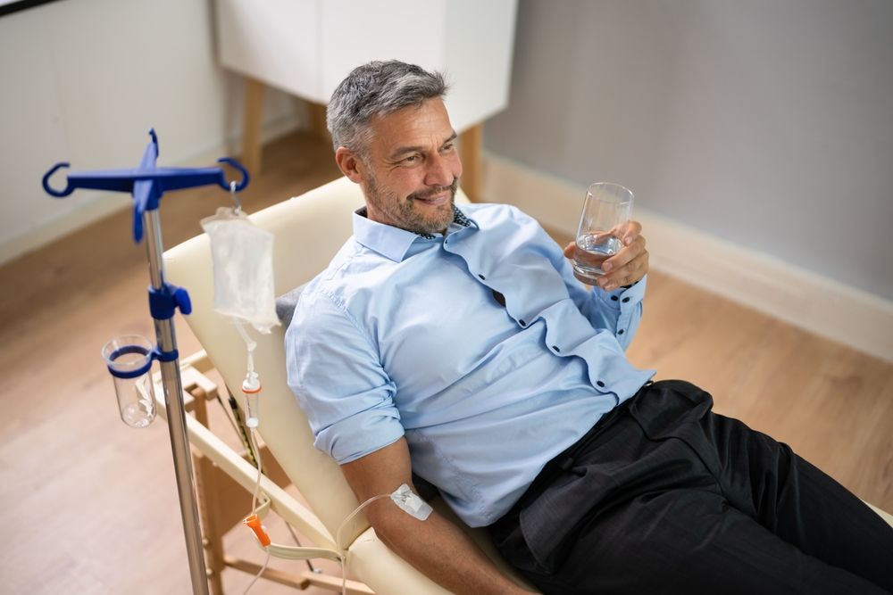 Man reclining in a chair holding a glass, connected to medical tubing in a home setting