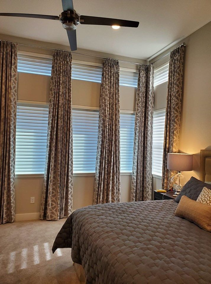 A bedroom with a large bed , a ceiling fan , and printed window curtains.