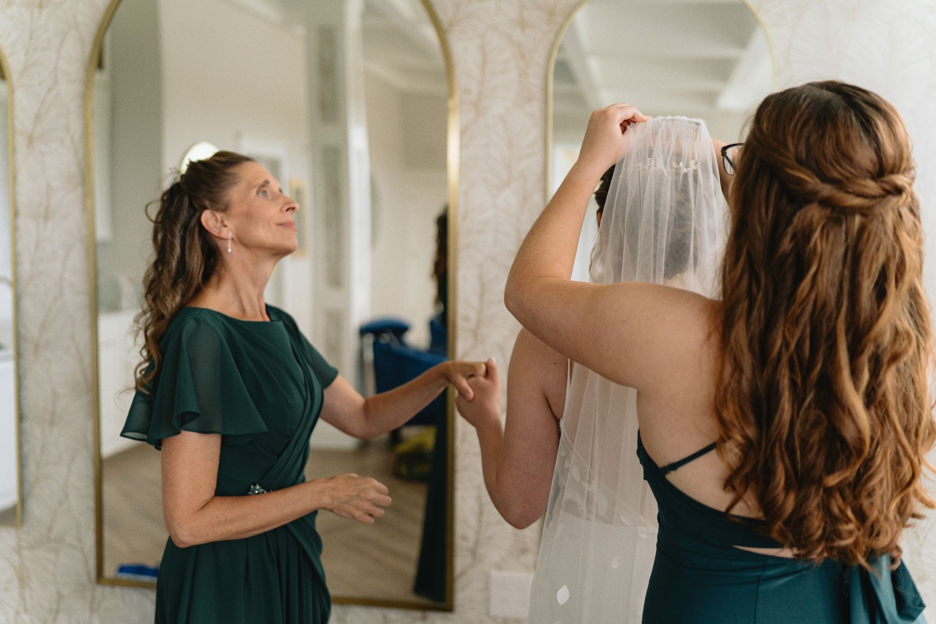 A bride and her mother are putting on a veil in front of a mirror.