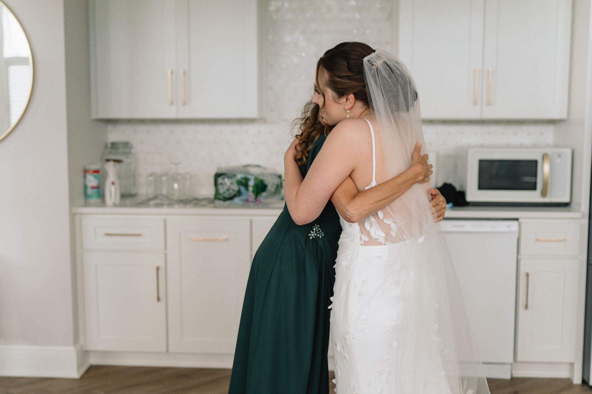 A bride and her bridesmaid are hugging each other in a kitchen.