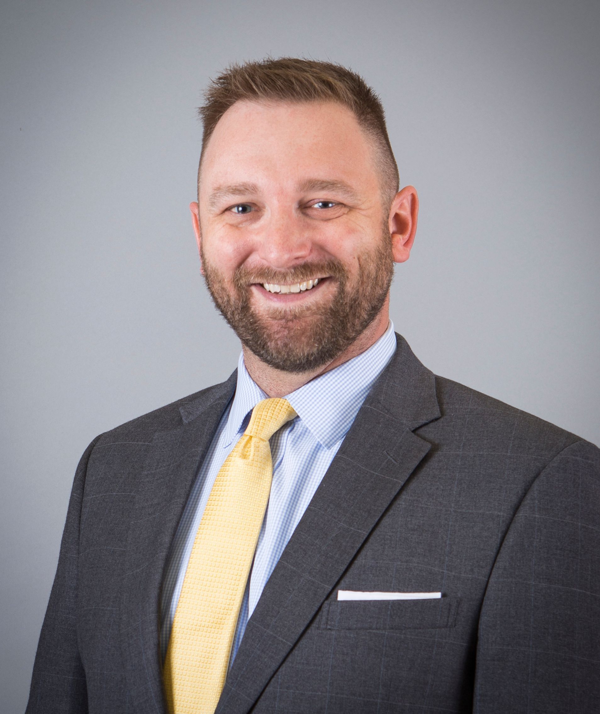 Man in gray suit with yellow tie smiles, looking towards the viewer. Light blue shirt, gray background.