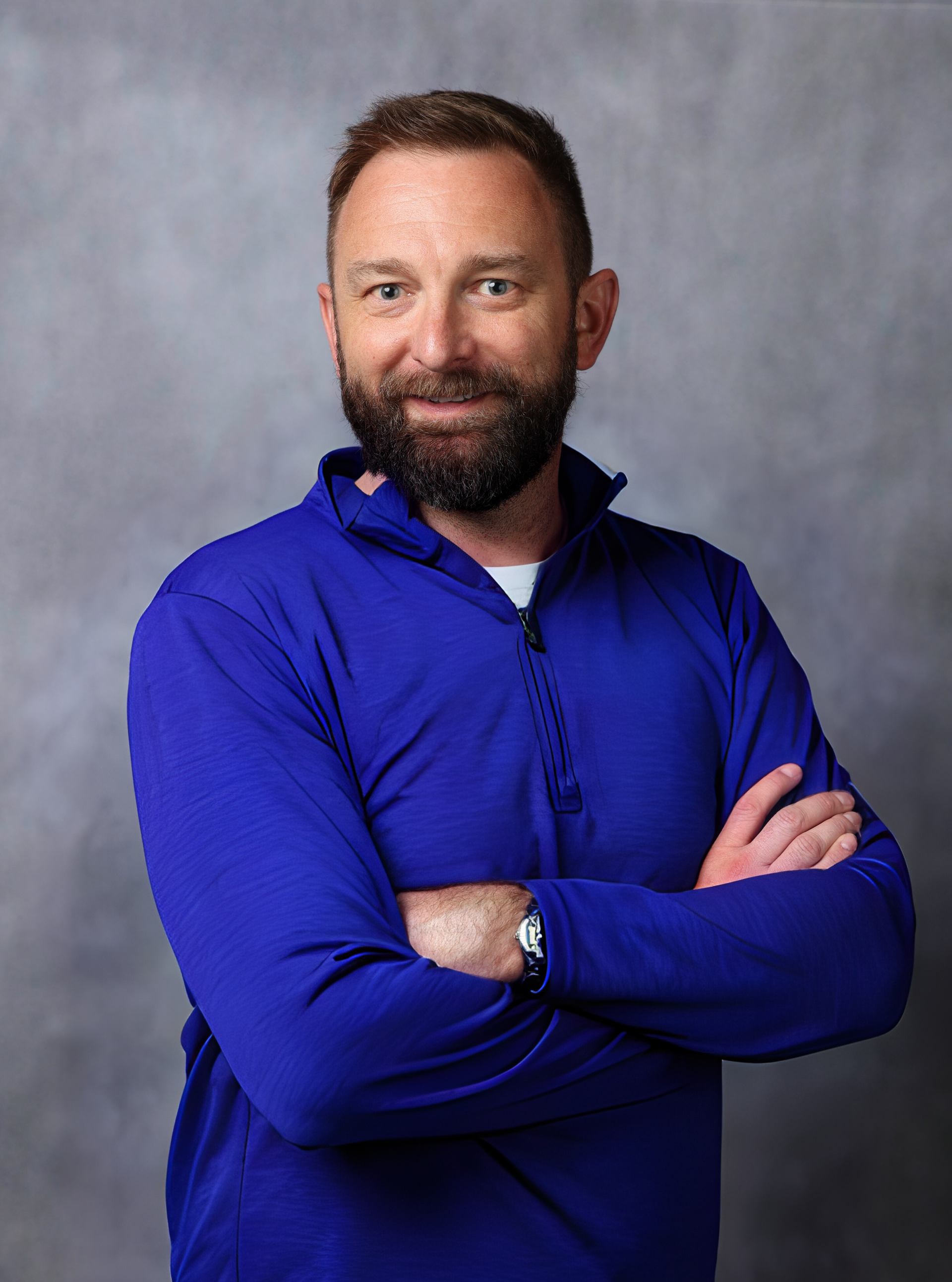 Man with beard and crossed arms, wearing a blue zip-up shirt, smiles against a grey backdrop.