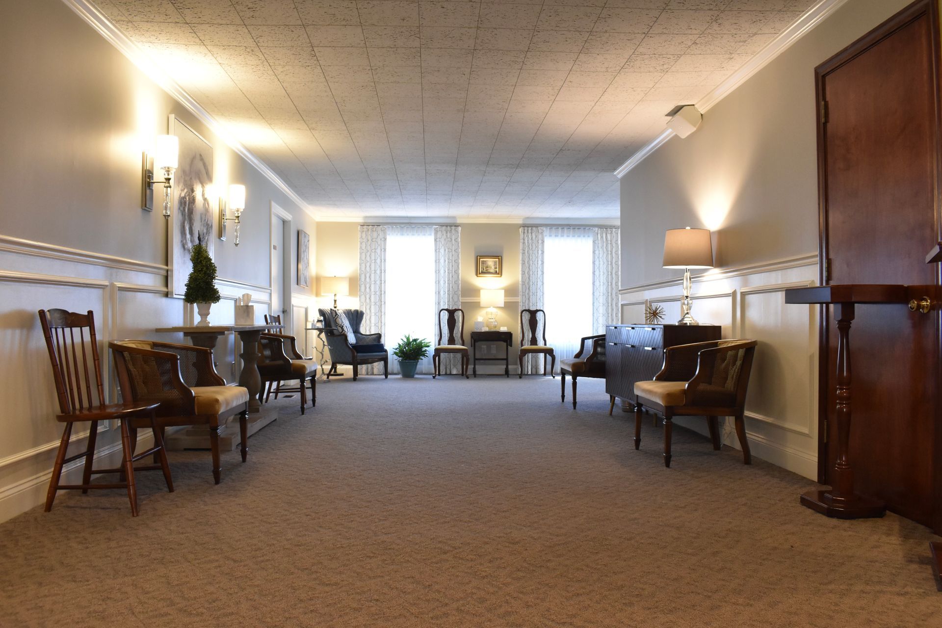 A long hallway with chairs , tables , lamps and a fireplace.