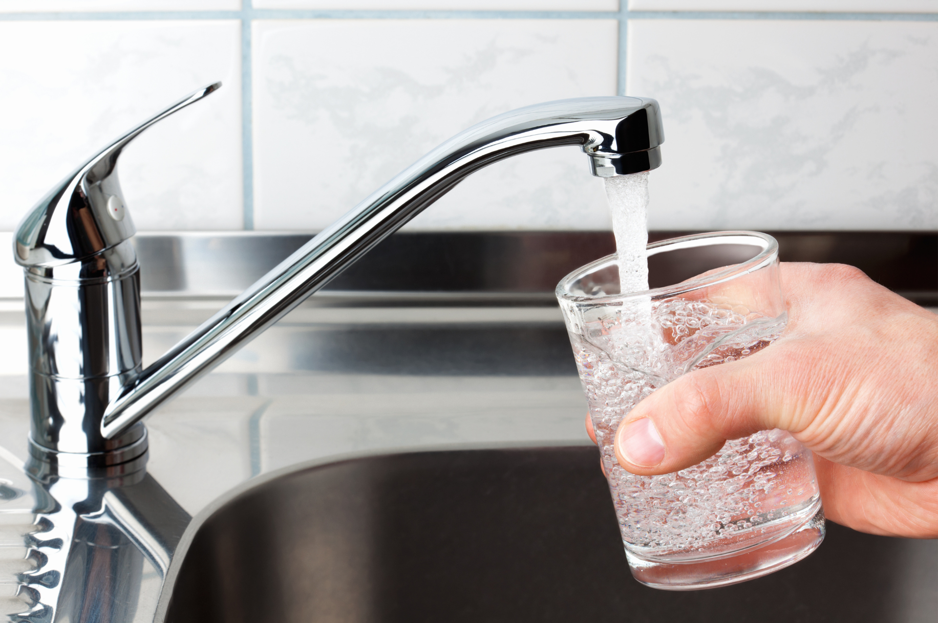 A person fills a glass with water from a chrome faucet in a kitchen sink.