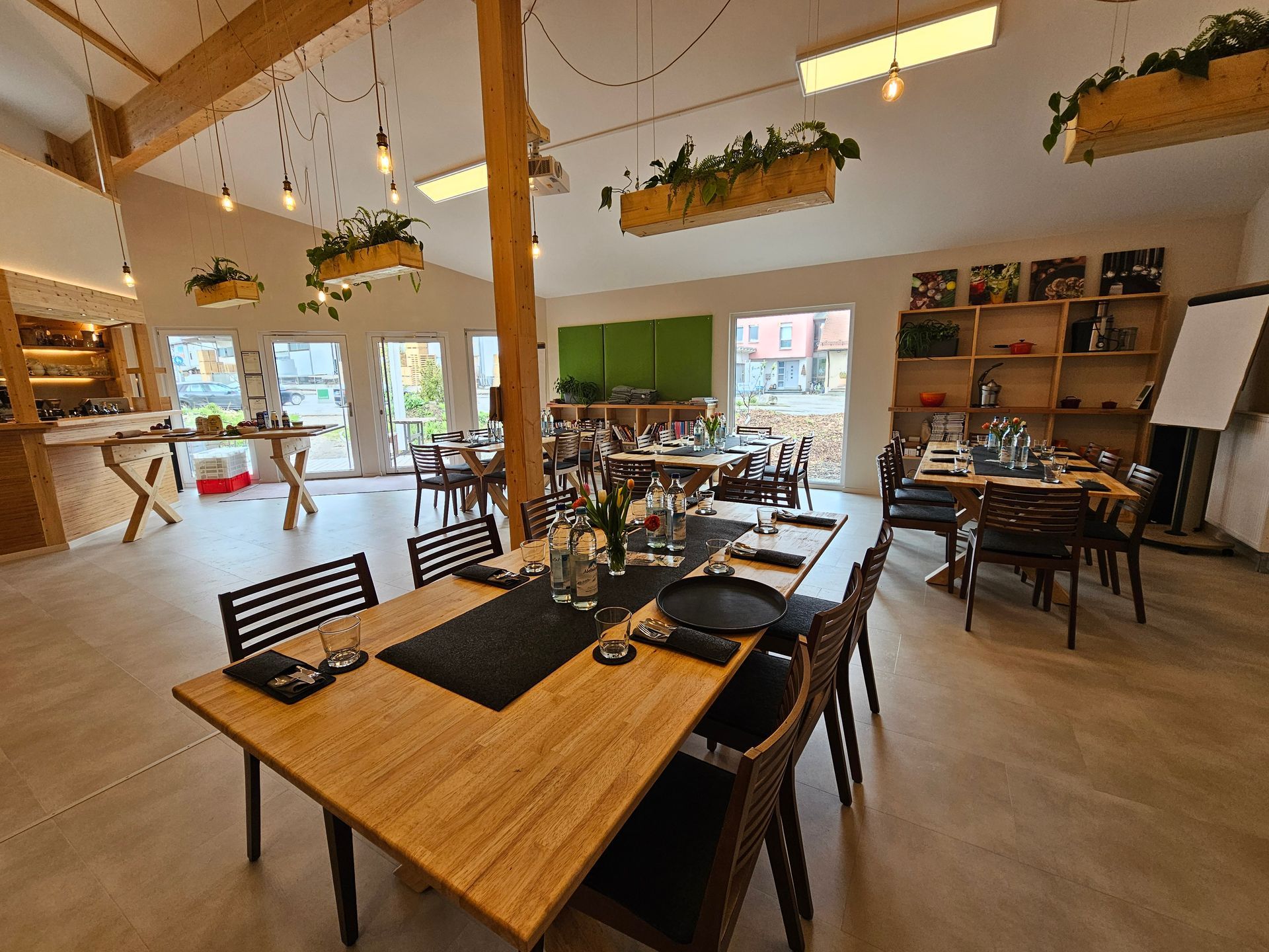 Innenraum eines hellen Raumes mit Holztischen, Stühlen und Hängepflanzen. Die Tische sind für eine Mahlzeit gedeckt.