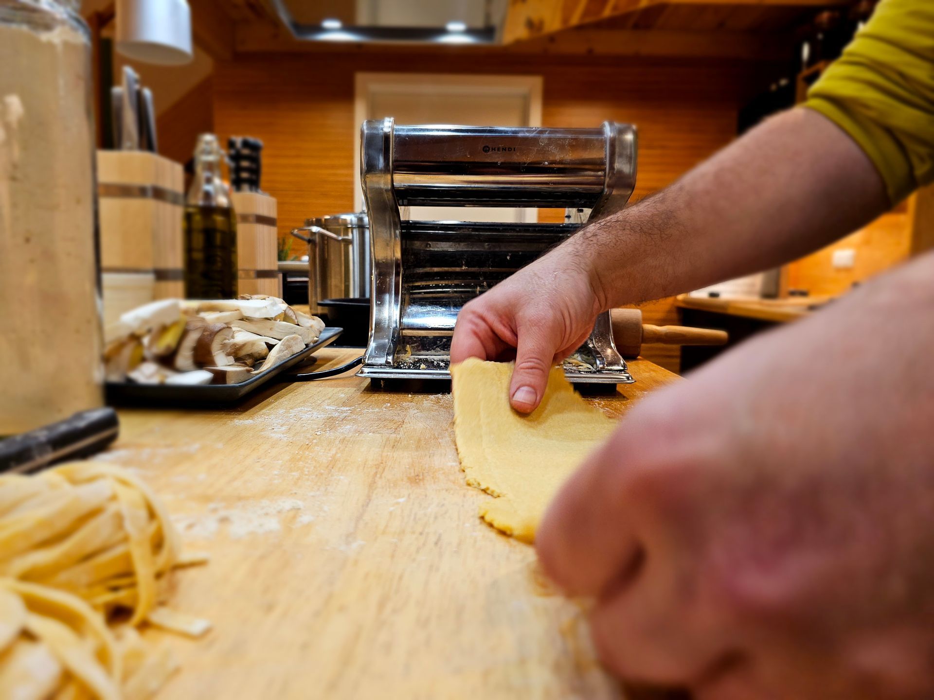 Person, die Nudelteig durch eine Maschine auf einem Holztisch rollt. Nudeln und Zutaten sind sichtbar.
