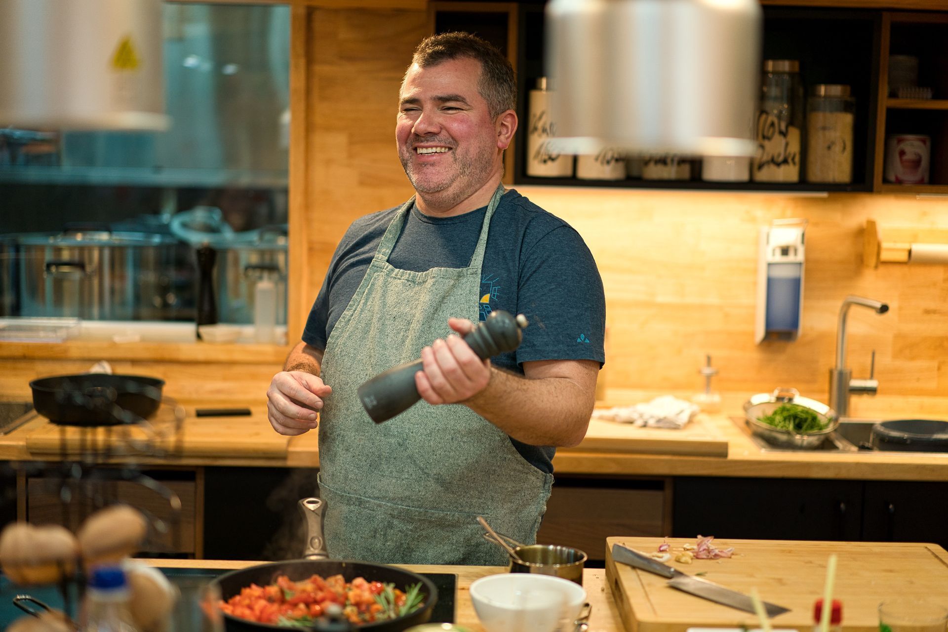 Lächelnder Koch in der Küche, Pfeffermühle in der Hand, Tomaten in einer Bratpfanne auf der Arbeitsplatte.
